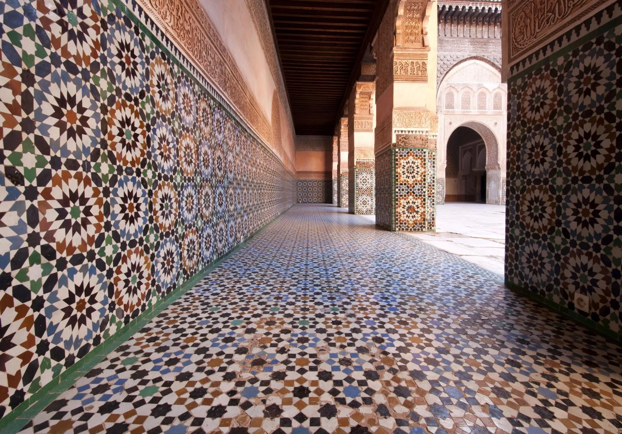Galerie de la médersa Ben Youssef aux murs couverts de zellige multicolore, avec des stucs sculptés au-dessus et la cour en marbre au fond